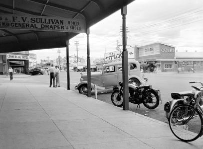 View down a wide street from under a wide verandah with parked cars, motorcycles and bicycle.