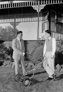 2 well-dressed men standing on a lawn beside a house. One of the men holds a push lawn mower.