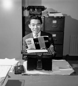 Portrait of a young man looking toward the camera while seated at a typewriter.