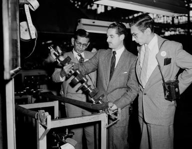 3 men in suits, in an engineering factory inspect a crankshaft.