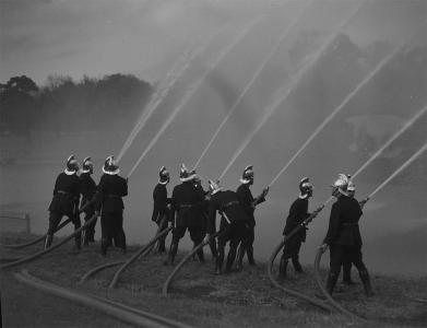 Firemen wearing shiny helmets practicing with fire hoses.