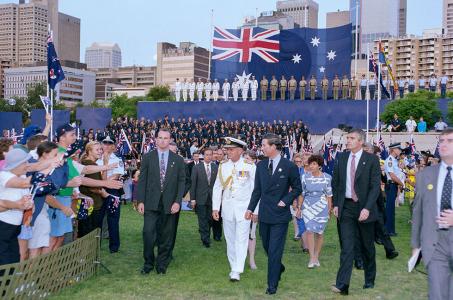 Prince Charles greeting the crowd at Darling Harbour. In the background are Australian military personnel assembled on a raised platform before a large Australian flag.