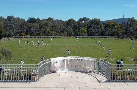 A cricket match on an oval. Hanging on a balcony in front of the match is a sign reading 'It's just not cricket. Let the Timorese stay.'