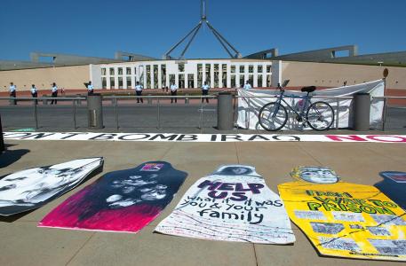 Protest signs lying on the ground outside Parliament House. They feature the faces of Iraqi people and handwritten text like 'Help us. What if it was your family.' and 'Don't bomb Iraq.'