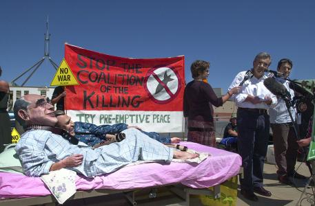 Protesters wearing masks of John Howard and George Bush lying in a bed in front of a sign saying 'Stop the coalition of killing. Try pre-emptive peace.' Parliament House is in the background. 