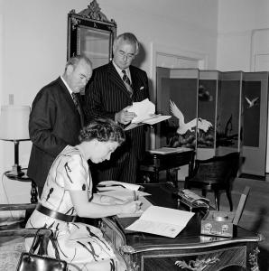 Paul Hasluck and Prime Minister Whitlam standing. Queen Elizabeth II seated at an ornate desk signing documents. An 8-panel screen depicting storks is visible in the background.