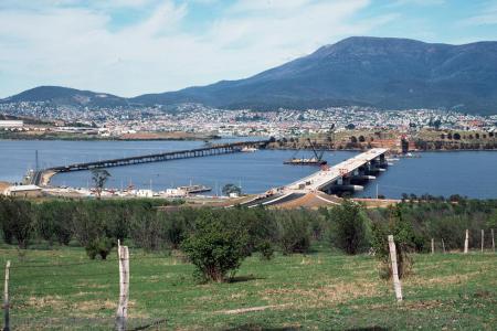 2 low bridges across a wide river. Small orchard in the foreground, suburban housing below a mountain in the background.